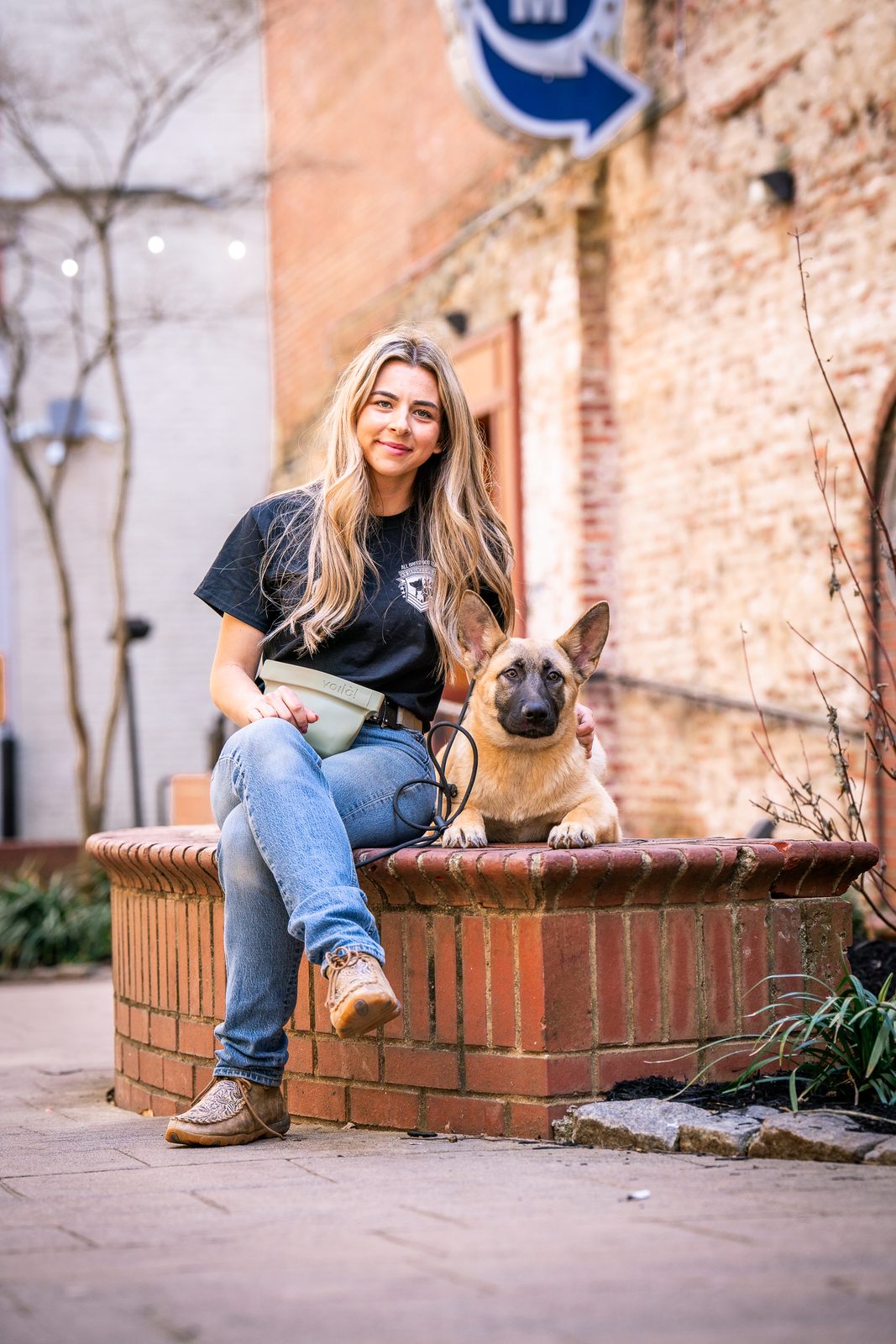 A woman sits on a brick planter with a young Belgian Malinois beside her