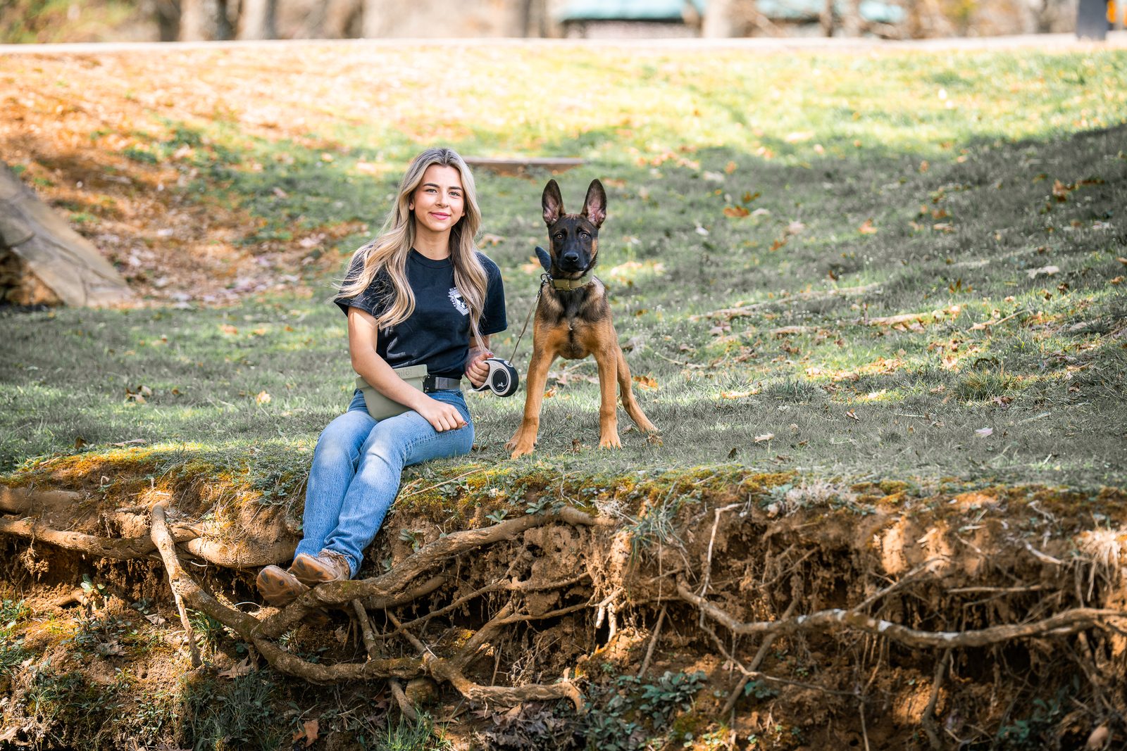 A woman sits on a mossy bank with a young Belgian Malinois beside her in warm autumn light